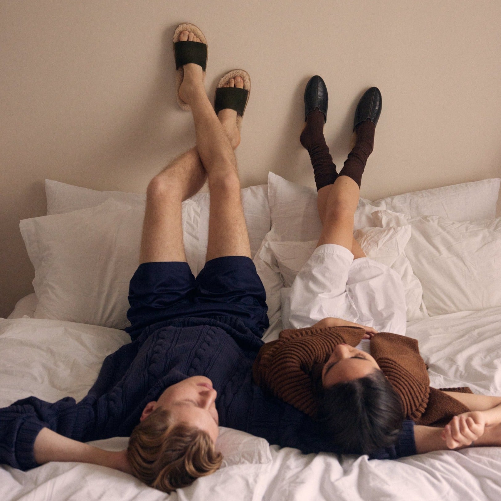One man and a women lying in bed with their feet up on the wall wearing Inabo slippers