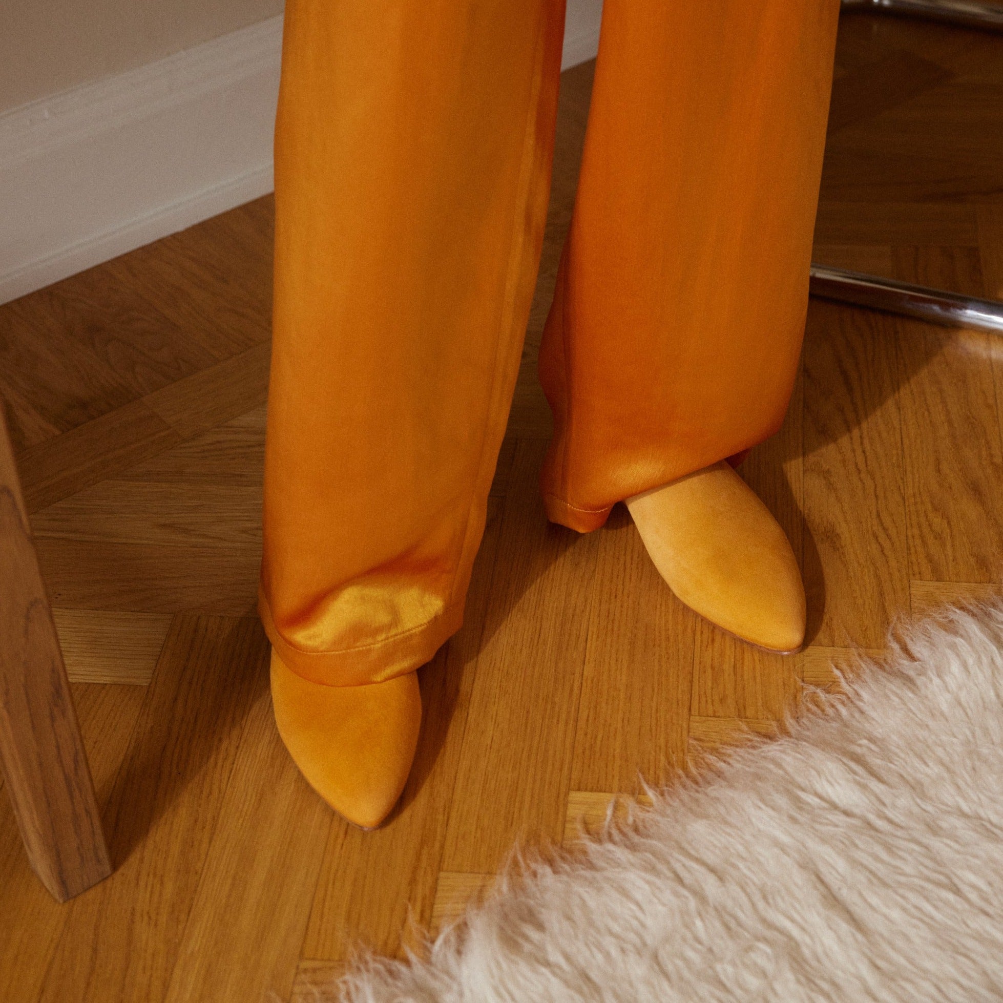 Inabo Women's Slider slipper in Saffron Suede worn by a women in orange satin pants standing on a wooden floor by a fluffy white rug