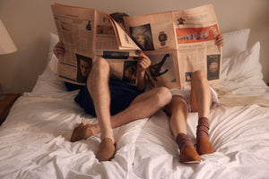 Man and Woman sitting on a bed reading a paper magazine with Brown suede slippers on their feet
