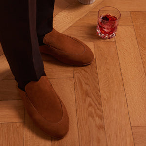 Inabo Men's Slowfer slipper in brown suede worn by a man standing on a wooden floor with a red drink 