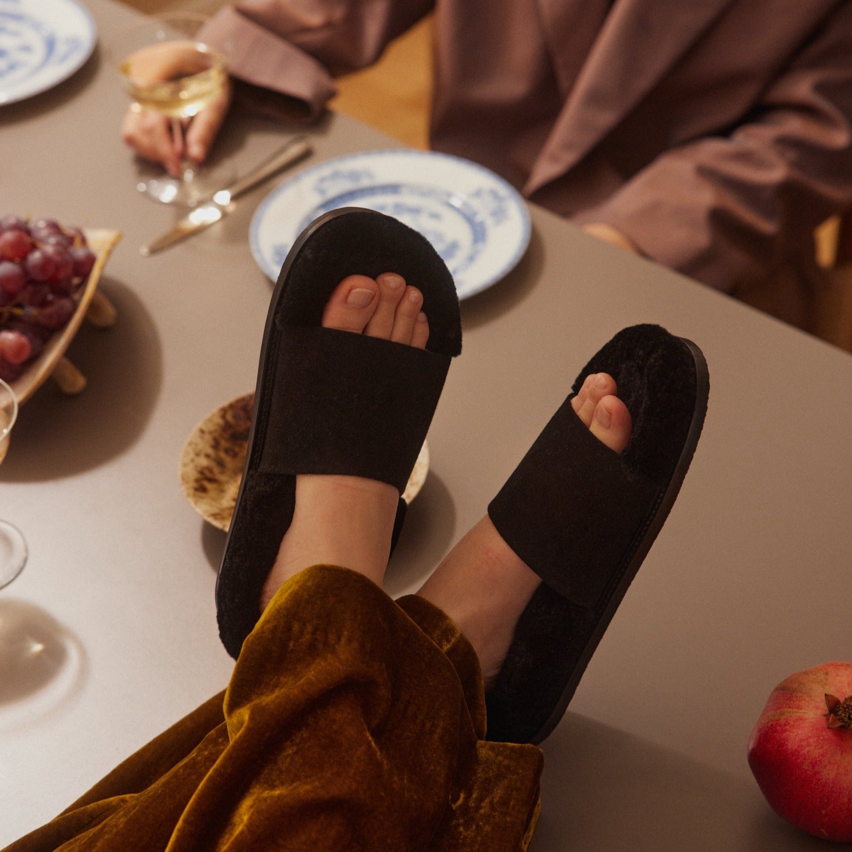 Inabo Women's Slipper in Black suede and shearling with an open-toe worn by a women sitting with her feet up on a laid table