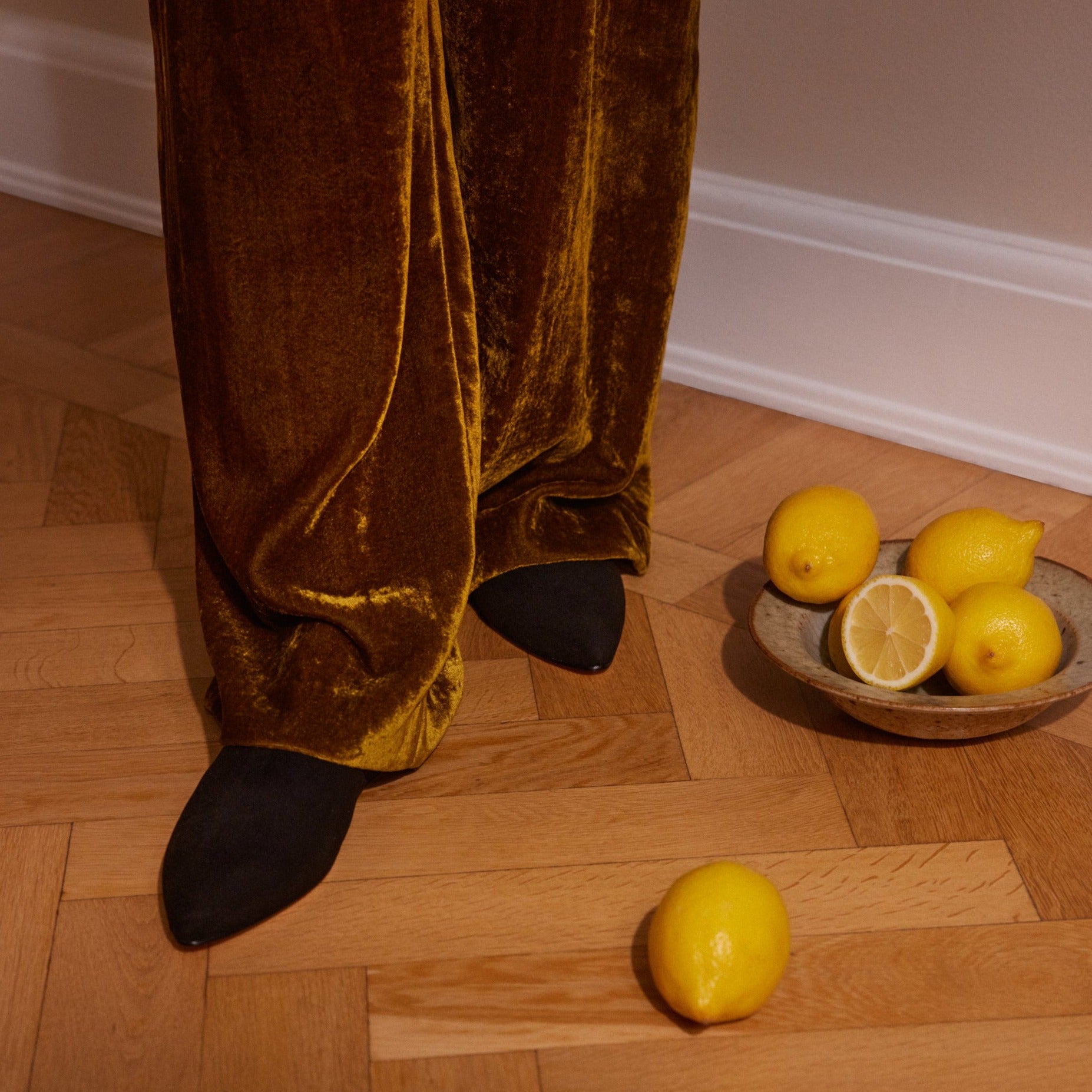 Inabo Women's Slider slipper in black suede worn by a women i velvet pants standing on a wooden floor with a bowl of lemons beside her 
