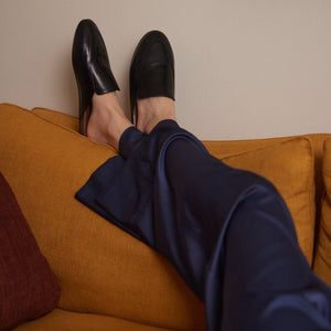 Inabo Women's Slowfer Slipper in black leather and brown leather insole worn by a woman in blue satin pants with her feet up on a yellow couch